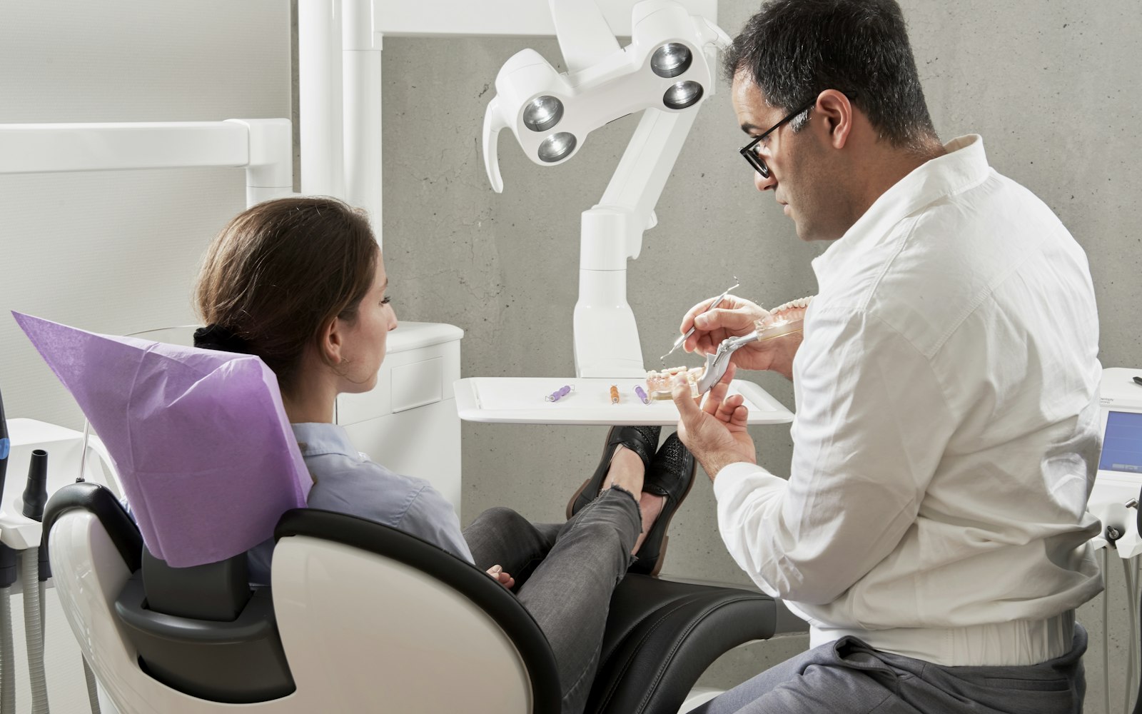 Dentist in a white shirt showing a model of teeth to a seated patient in a bright, modern treatment room
