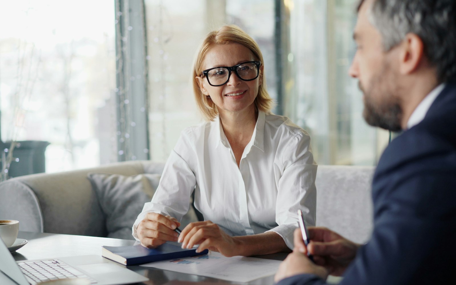 Woman in glasses and a white shirt listens to a man in a suit across a table with a notebook, papers and a laptop