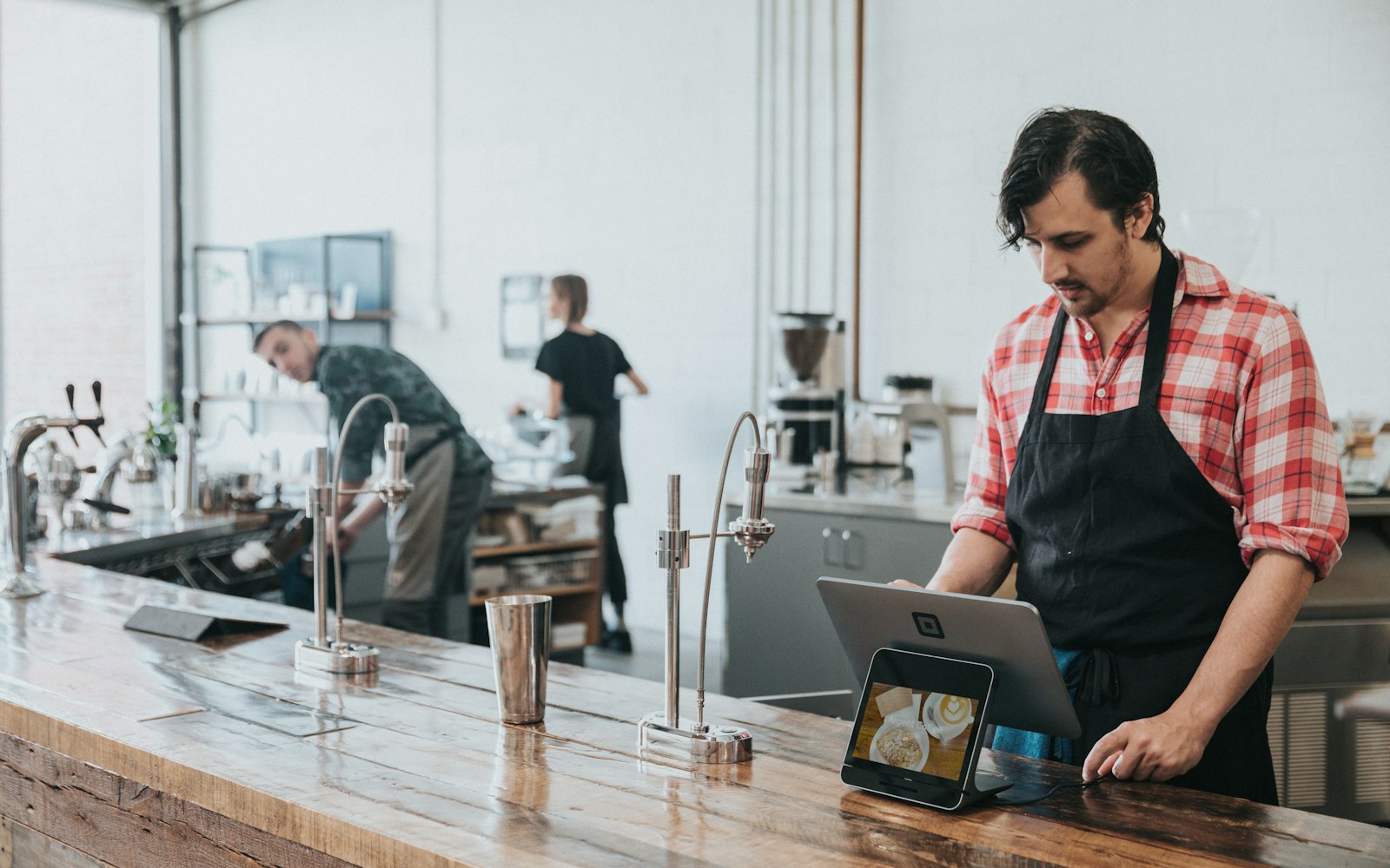 Café owner in a plaid shirt and apron checks a tablet at a wooden counter while staff work behind him