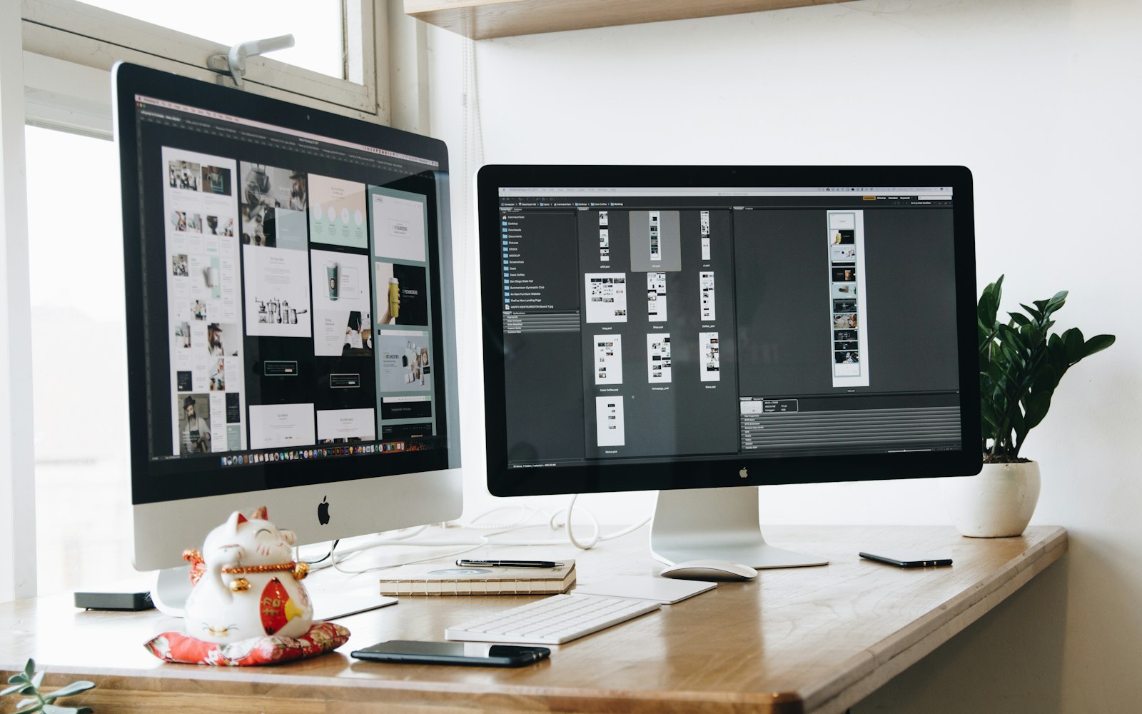 Two desktop monitors showing website page layouts on a wooden desk by a sunlit window, with a notebook, pen and plant